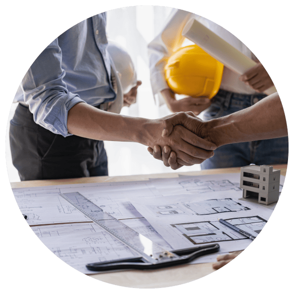 Three men holding hard hats are conversing over a desk with architectural plans and a model of a building; two of the men are shaking hands.