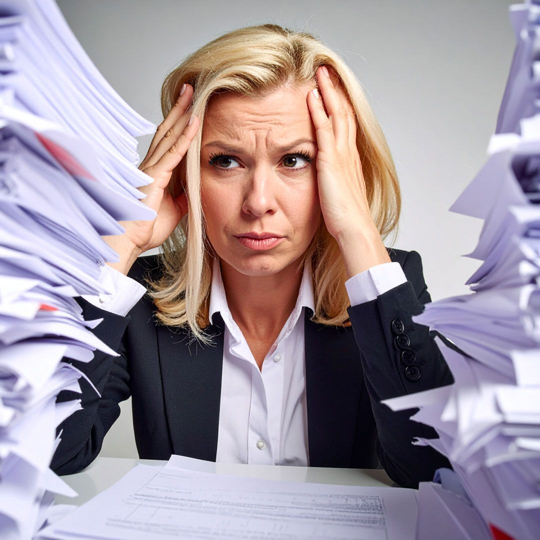 A woman sits at her desk with her hands on her head, surrounded by towering piles of paperwork, her face etched with exhaustion and frustration, clearly overwhelmed by the mounting pressure of her workload.