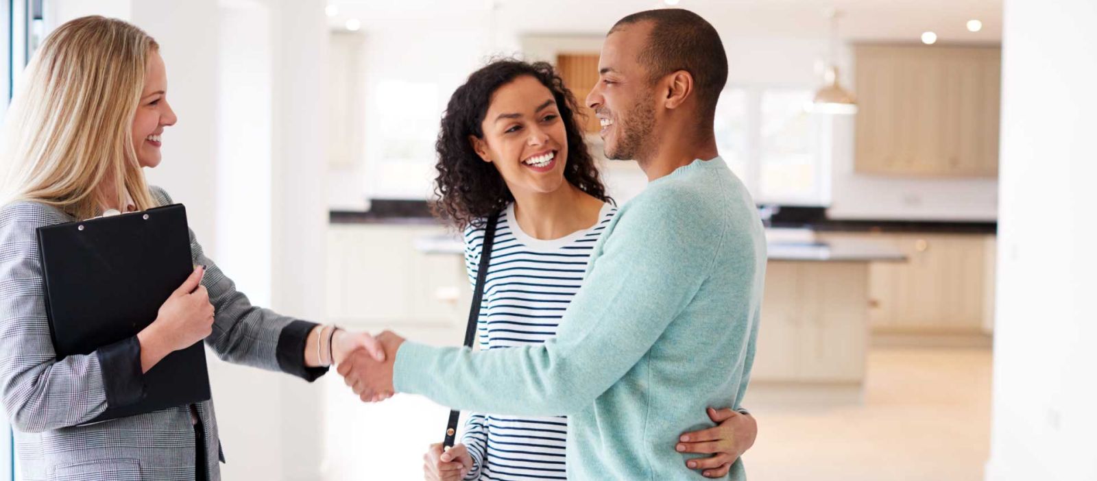 A female realtor holding a clipboard is shaking hands with a smiling couple in an empty well lit house.