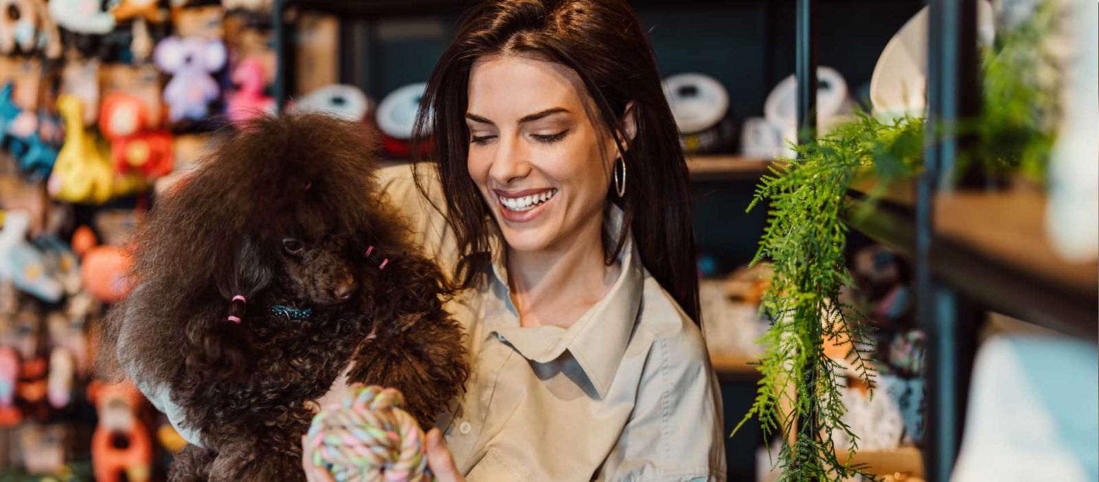 A smiling woman holds her dog while shopping in a pet store; there are various pet toys and supplies in the background.