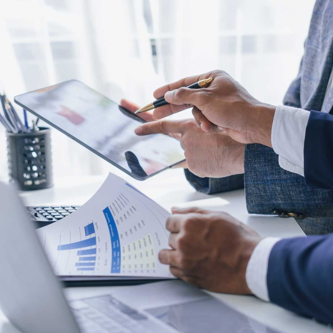 A man's hand holds a pen pointing at coworker's tablet while sitting on a desk with other documents displaying financial data.